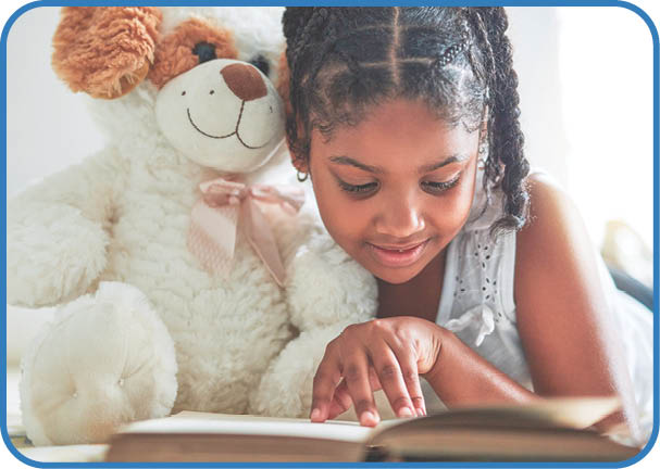 Shot of a young girl lying on bed with her teddybear while reading a book