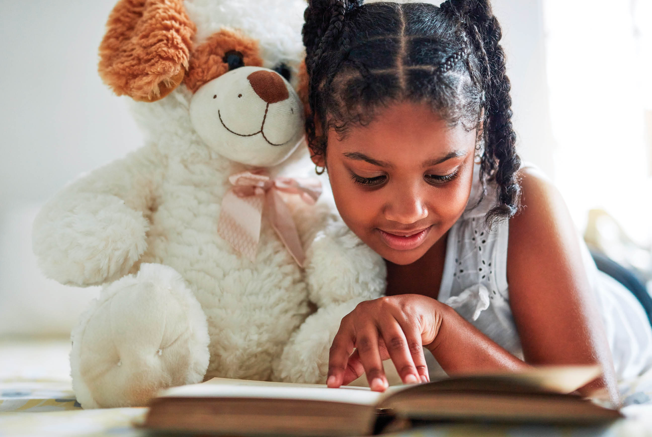 Shot of a young girl lying on bed with her teddybear while reading a book